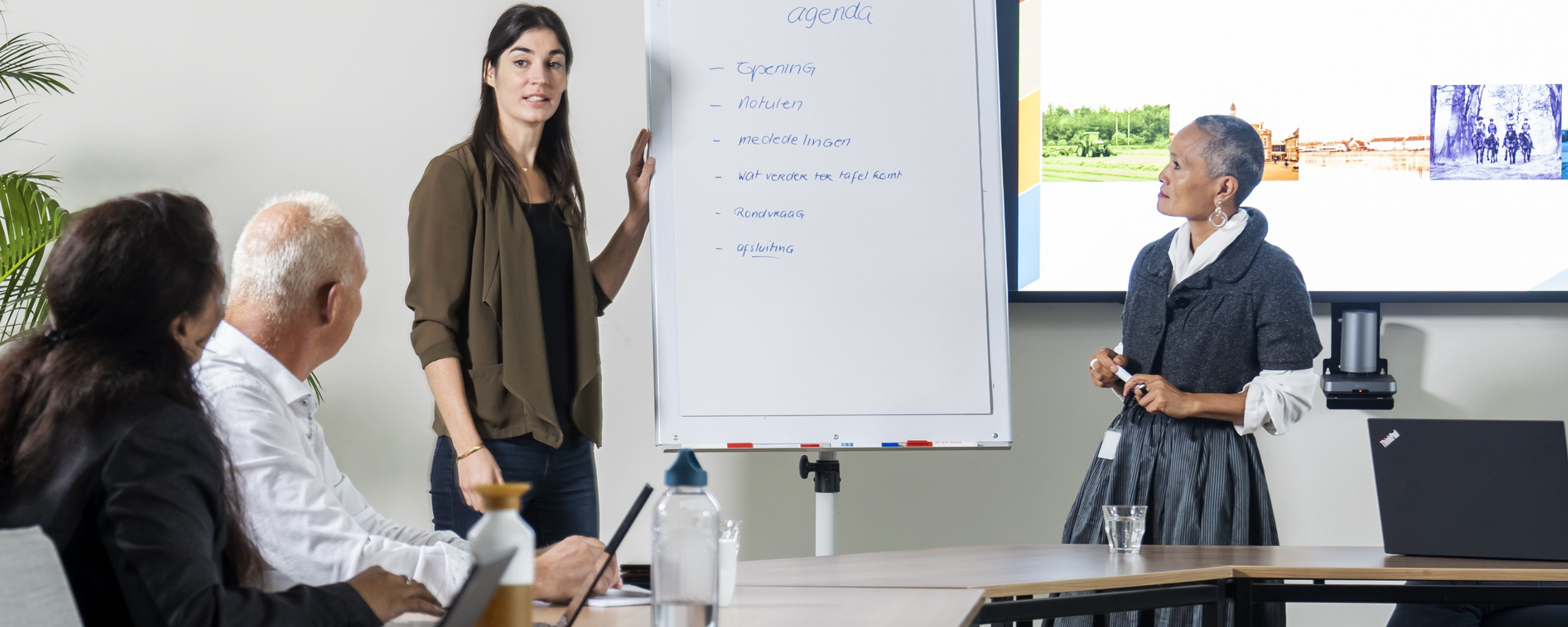 Een aantal medewerkers zit en staat in een vergadering en zij maken gebruik van het scherm en de flip-over.