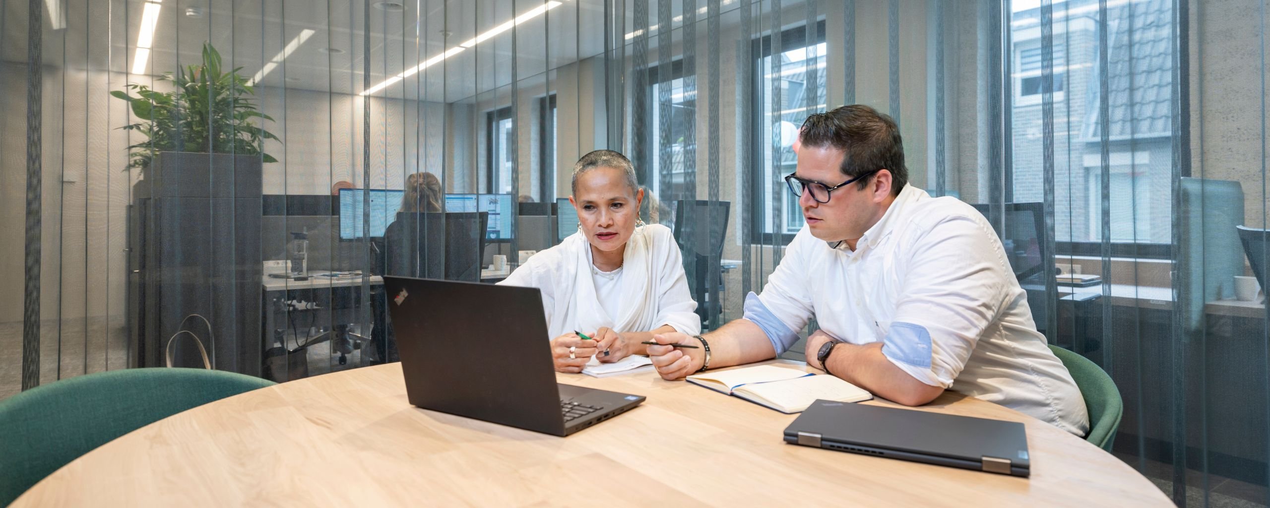 Twee medewerkers zitten in een vergadering te overleggen en aantekeningen te maken, kijkend naar het scherm van de laptop.