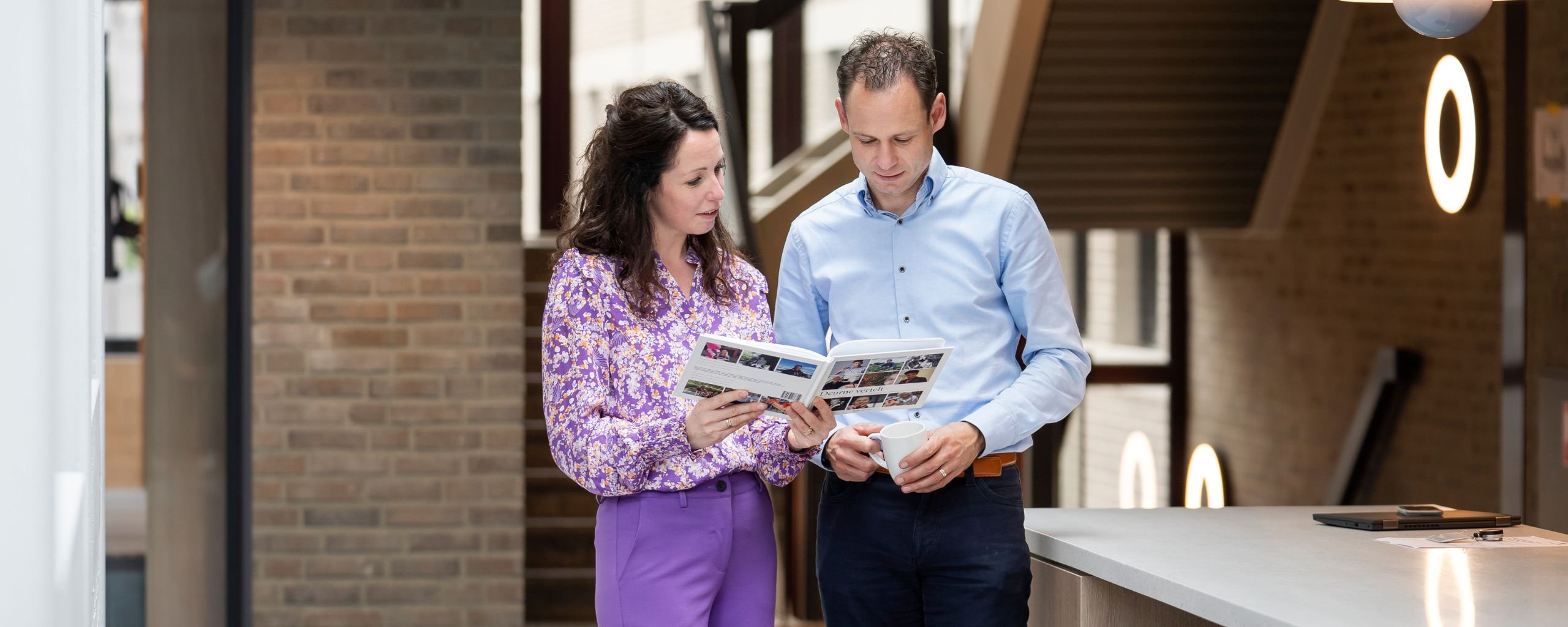Twee medewerkers staan in de hal en kijken in een boek waar ze het over hebben.