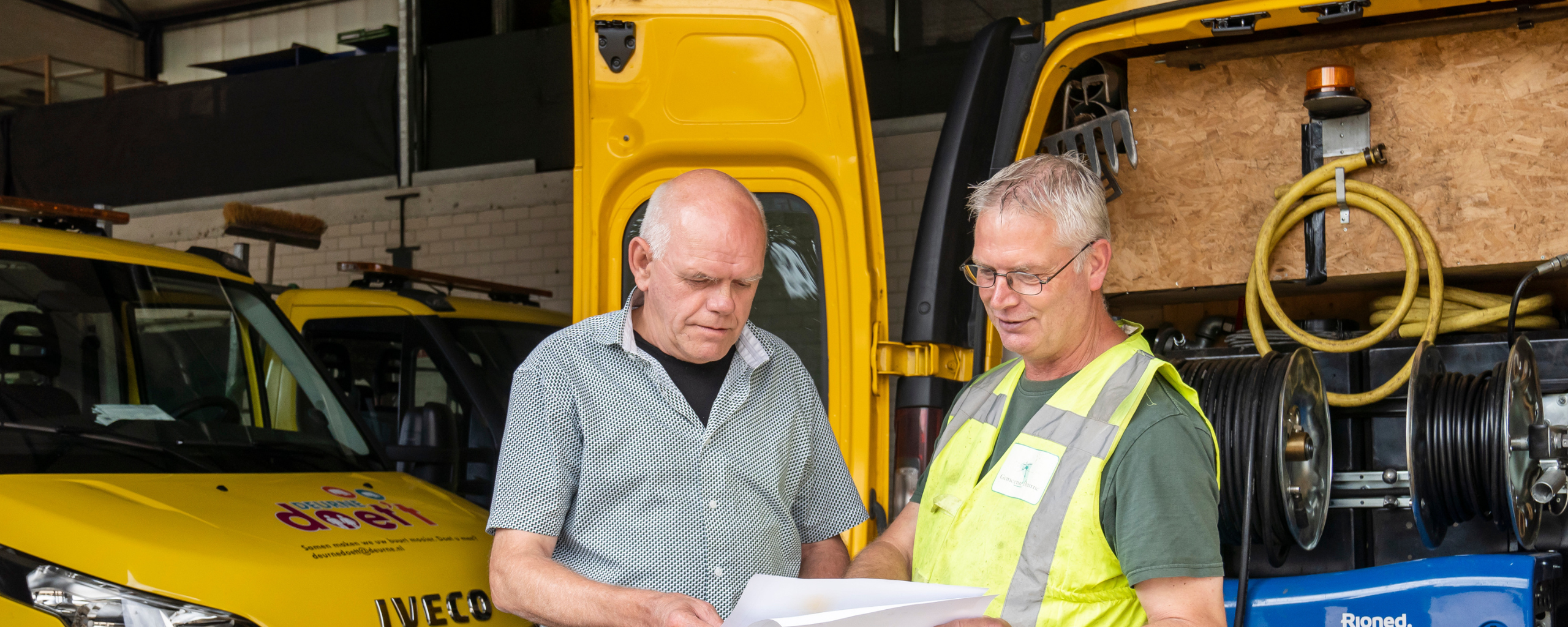 Twee buitendienst medewerkers bespreken samen wat er op het papier geschreven of afgebeeld staat.