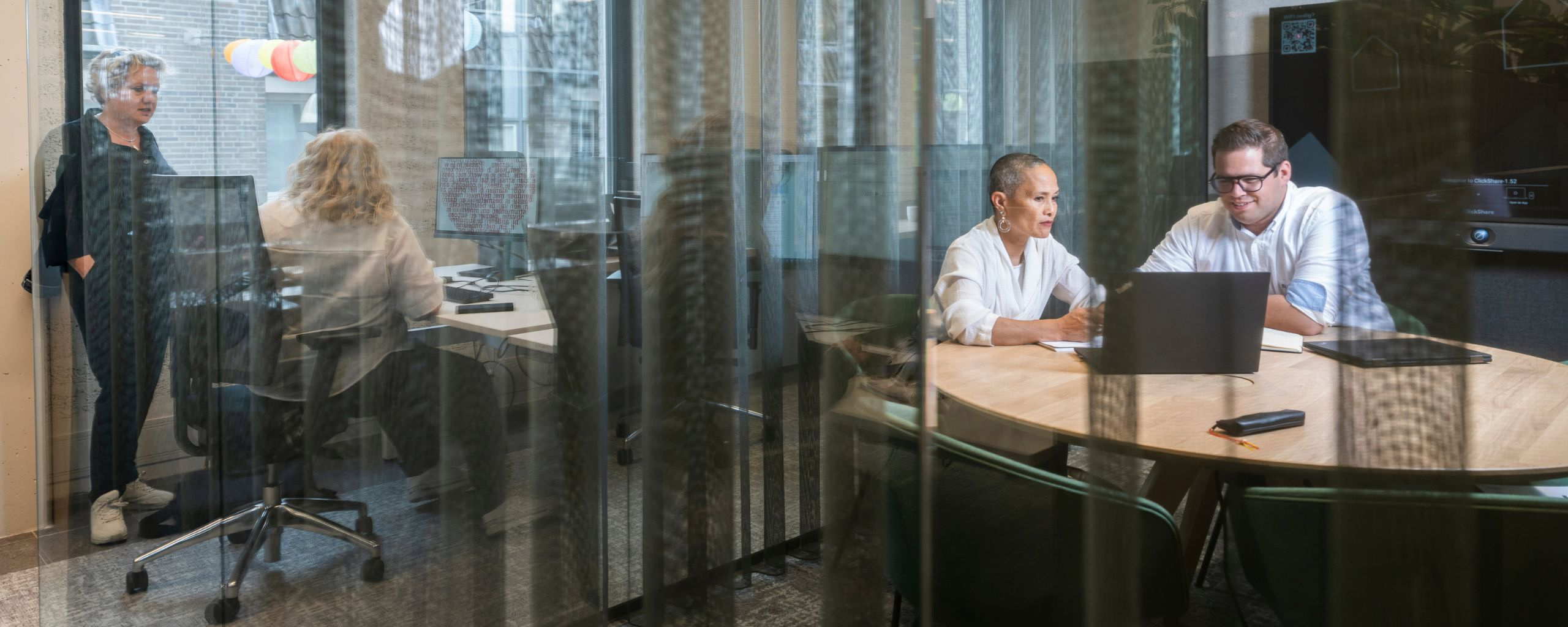 Er zijn 4 medewerkers te zien die in duo’s een gesprek voeren. Door het glas is het niet te verstaan waar de andere medewerkers het over hebben. Zo zit je dicht bij je collega’s, maar heb je toch de stilte die je op dat moment nodig hebt.