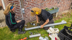 Twee vrouwen zijn aan het werk aan een afvoerpijp van regenwater