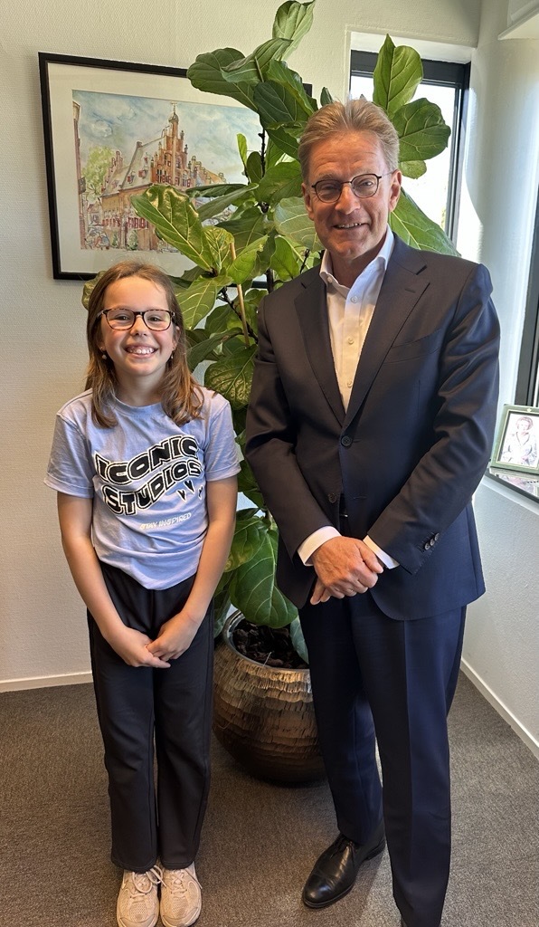Kinderburgemeester Emma naast burgemeester Bats, staand lachend, groene plant op achtergrond en vensterraam