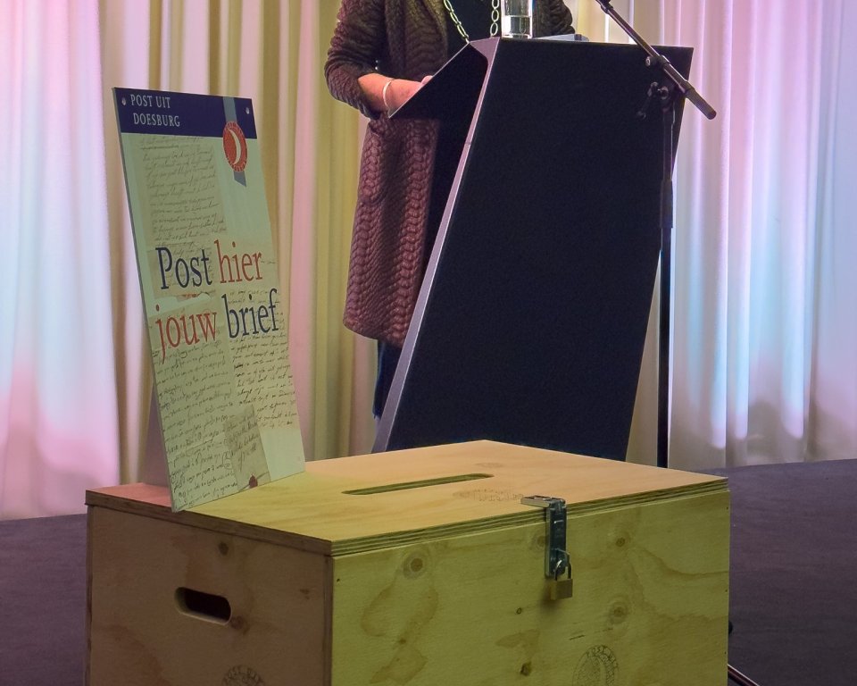 Vrouw (burgemeester) staand, hangende gordijnen, op voorgrond houten ideeënkistje met opschrift 'Post hier jouw brief' 