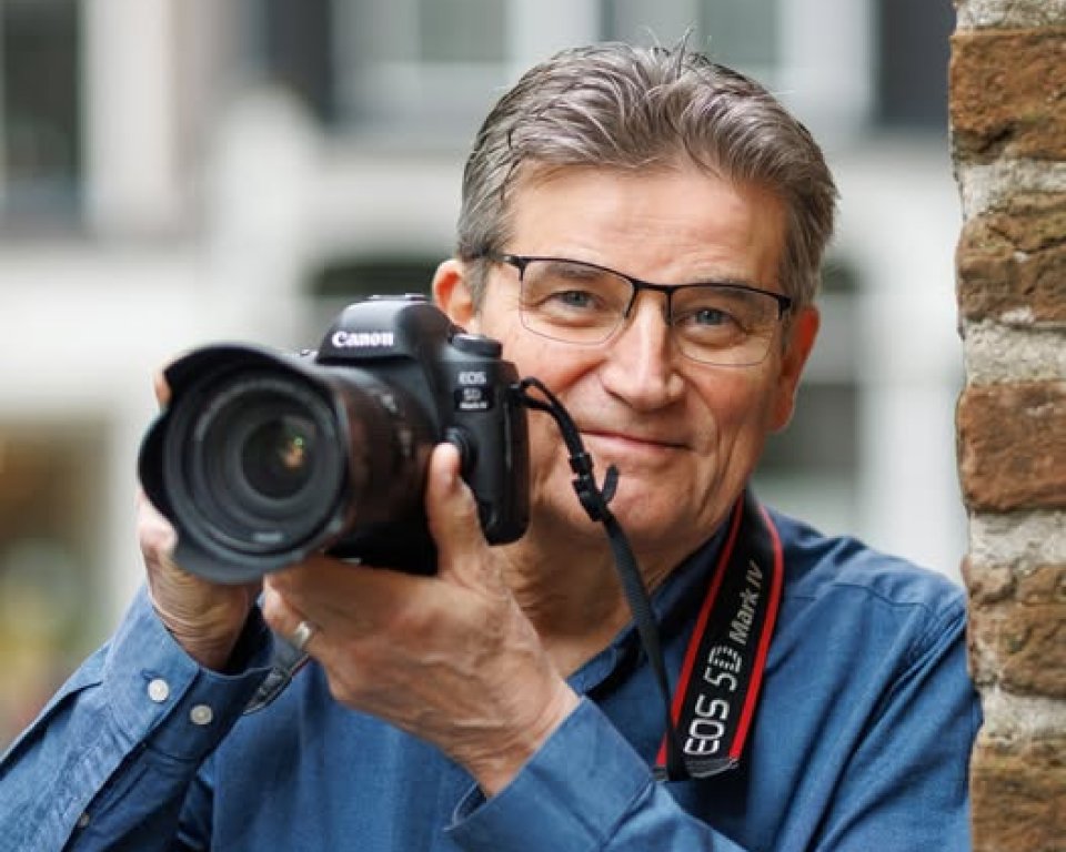 Man met bril en fotocamera in handen, lachend, leunend tegen muur