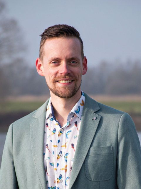 Man, lachend met baardje, licht fleurig overhemd en mintgroene pakjas en rivier, gras en bomen wazig op achtergrond