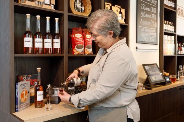 Eigenaar Granos koffiebar, vrouw staand, schenkt een glaasje Granos likeur in aan achterzijde toonbank
