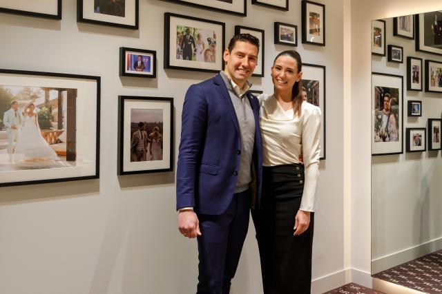 Man en vrouw staande naast elkaar lachend, ingelijste portretten op de achtergrond en grote spiegel rechts