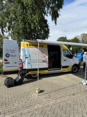 Op de afbbeelding een witte bestelbus met gele strepen, mensen op de achtergrond, een boom