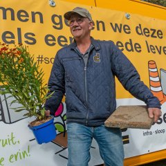 Man lachend rechtopstaand met in de linkerhand een tuintegel en in de rechterhand een tuinplant