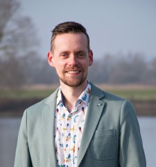 Man, lachend met baardje, licht fleurig overhemd en mintgroene pakjas en rivier, gras en bomen wazig op achtergrond