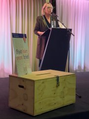 Vrouw (burgemeester) staand, hangende gordijnen, op voorgrond houten ideeënkistje met opschrift 'Post hier jouw brief' 