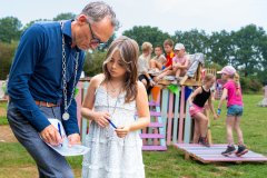Wethouder bij kind schrijft met pen op papier (map), staand buiten, kinderen op achtergrond