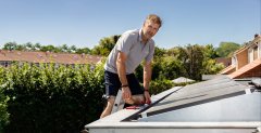 Man bevestigt zonneppanelen op dak van woning, achtegrond bomen, groen en rijtjeshuizen