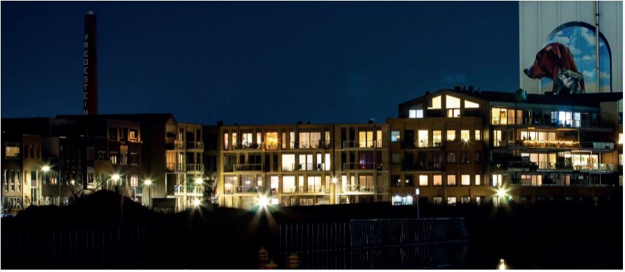 appartementencomplex op een donkere avond. in de huizen schijnt licht. Op de achtergond tegen de donkere lucht de schoorsteen van Vredestein. Rechts een grote silo met een tekening van een hond.