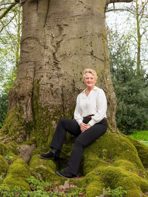 Georgette zit op de wortel van een ontzettend grote boom. Alleen de dikke stam is zichtbaar.  De wortels zijn begroeid met mos.