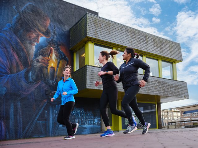 3 dames rennen op het plein voor het stadhuis. Op de achtergrond de muurschildering van de lantaarnaansteker.