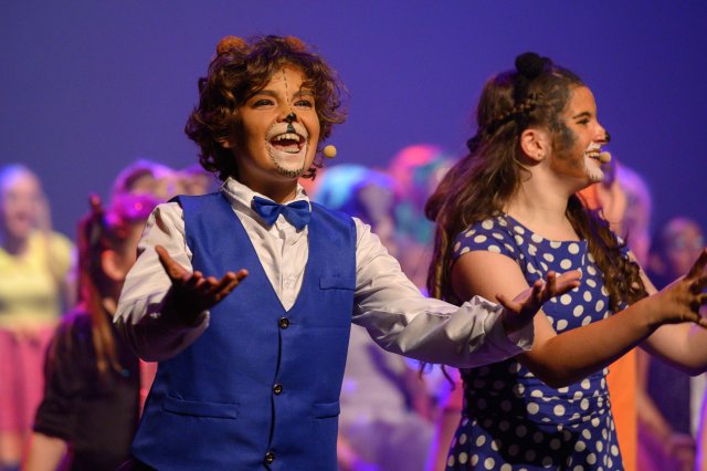 Eindmanifestatie van de musicalschool met een jongen en meisje op de voorgrond. De jongen is om de mond geschminkt. Ze hebben een lach op hun gezicht.