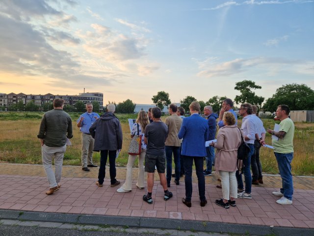 Een groep mensen staat te luisteren naar een man. Sommige personen hebben papieren in hun handen. Ze staan allemaal op een stoep voor een braakliggend terrein. Op de horizon staan enkel stadshuizen en bomen.