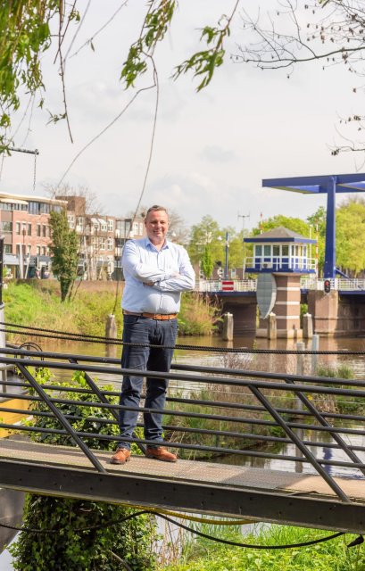 Gerben staat op een loopbrug voor het water. Op de achtergrond een ophaalbrug met een wachtershuisje. Aan de overkant van het water staan enkele appartementen.