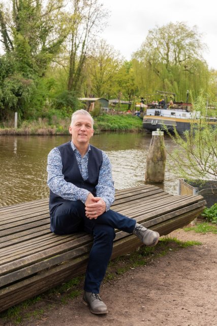 Hans zit op een bankje zonder rugleuning voor het water. Op de achtergrond ligt in het water een schip