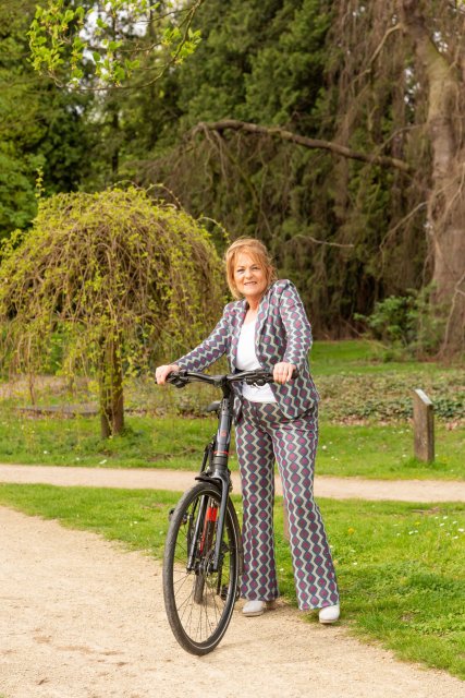 Marion staat met een fiets in haar handen. Op de achtergrond zijn bomen.