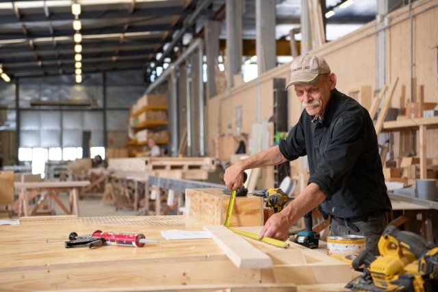 Man met pet aan het werk met hout in een grote hal.