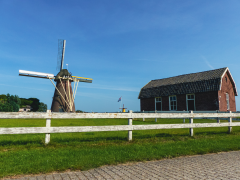 Op de voorgrond een wit houten hekwerk met daarachter grasland, rechts een boerderij en links een molen.