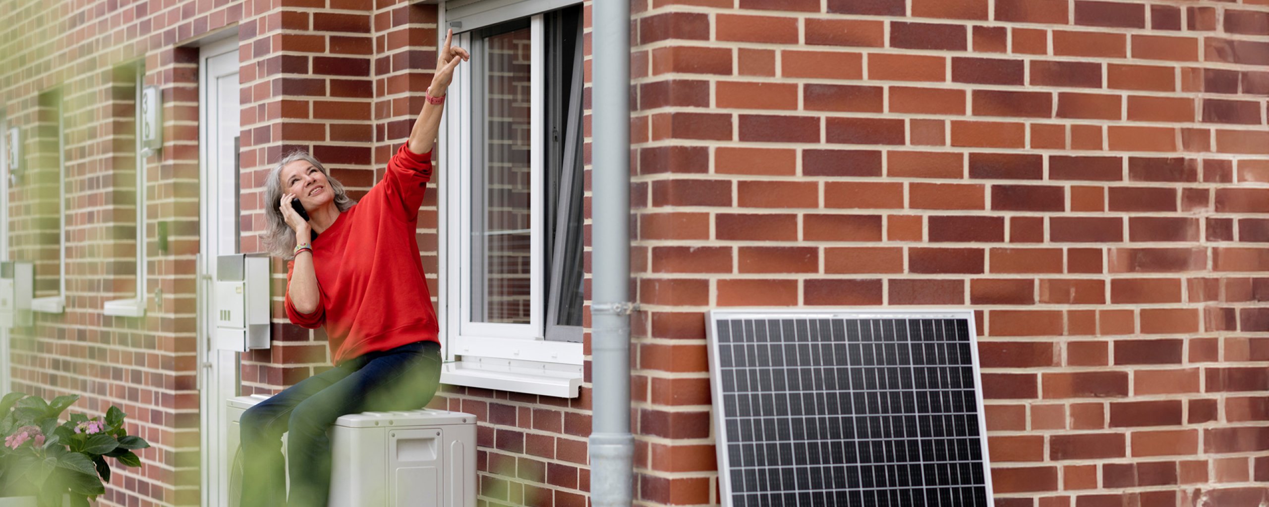 Vrouw met een rode trui, zittend op een warmtepomp voor een huis, met tegen een muur staand een zonnepaneel
