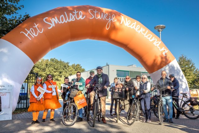 Deelnemers naast hun fiets onder de oranje boog van Het Smalste Stukje Nederland
