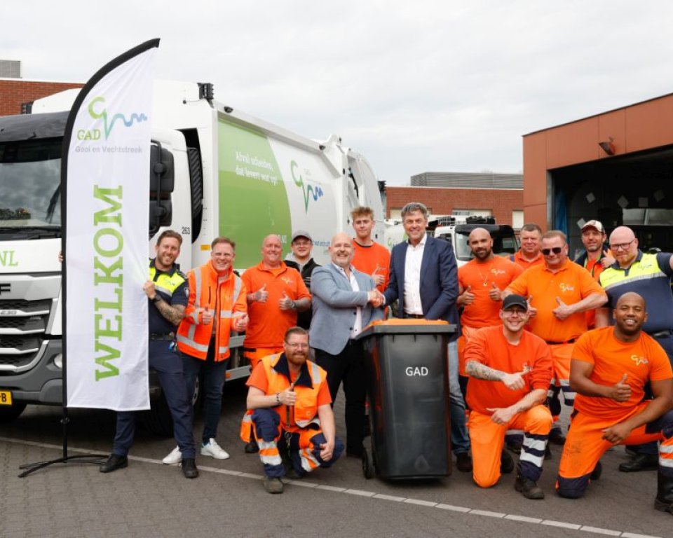 Medewerkers in felle kleding poseren met de wethouder voor een vuilniswagen op het terrein van GAD in Bussum
