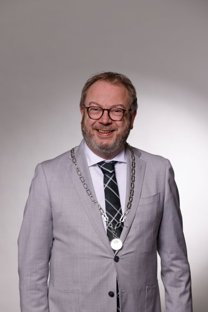 Persoon in een donker pak met ambtsketen, staand voor een lichte achtergrond.