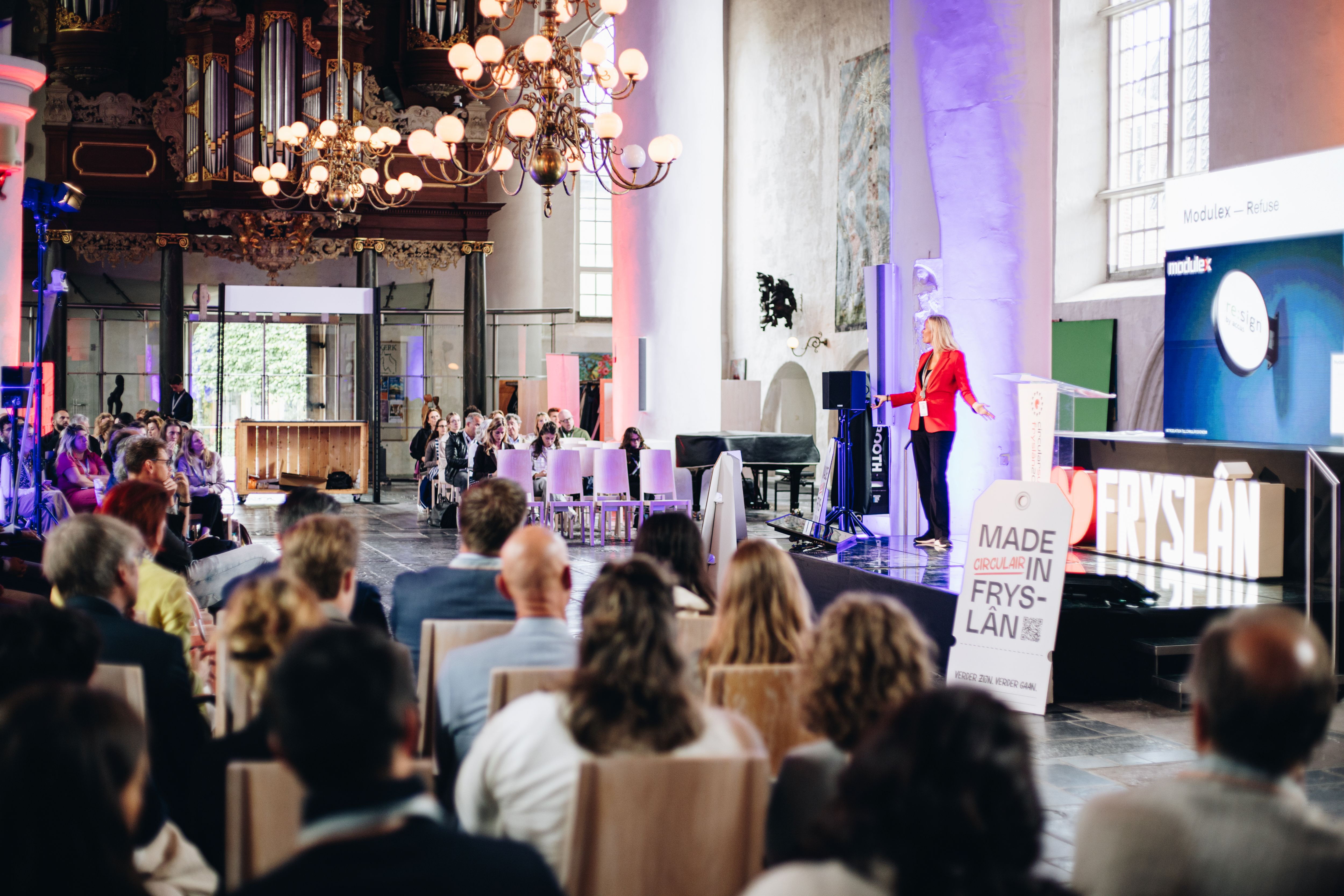 Een zaal vol publiek luisteren aandachtig naar een vrouw in een rood jasje die op het podium staat.