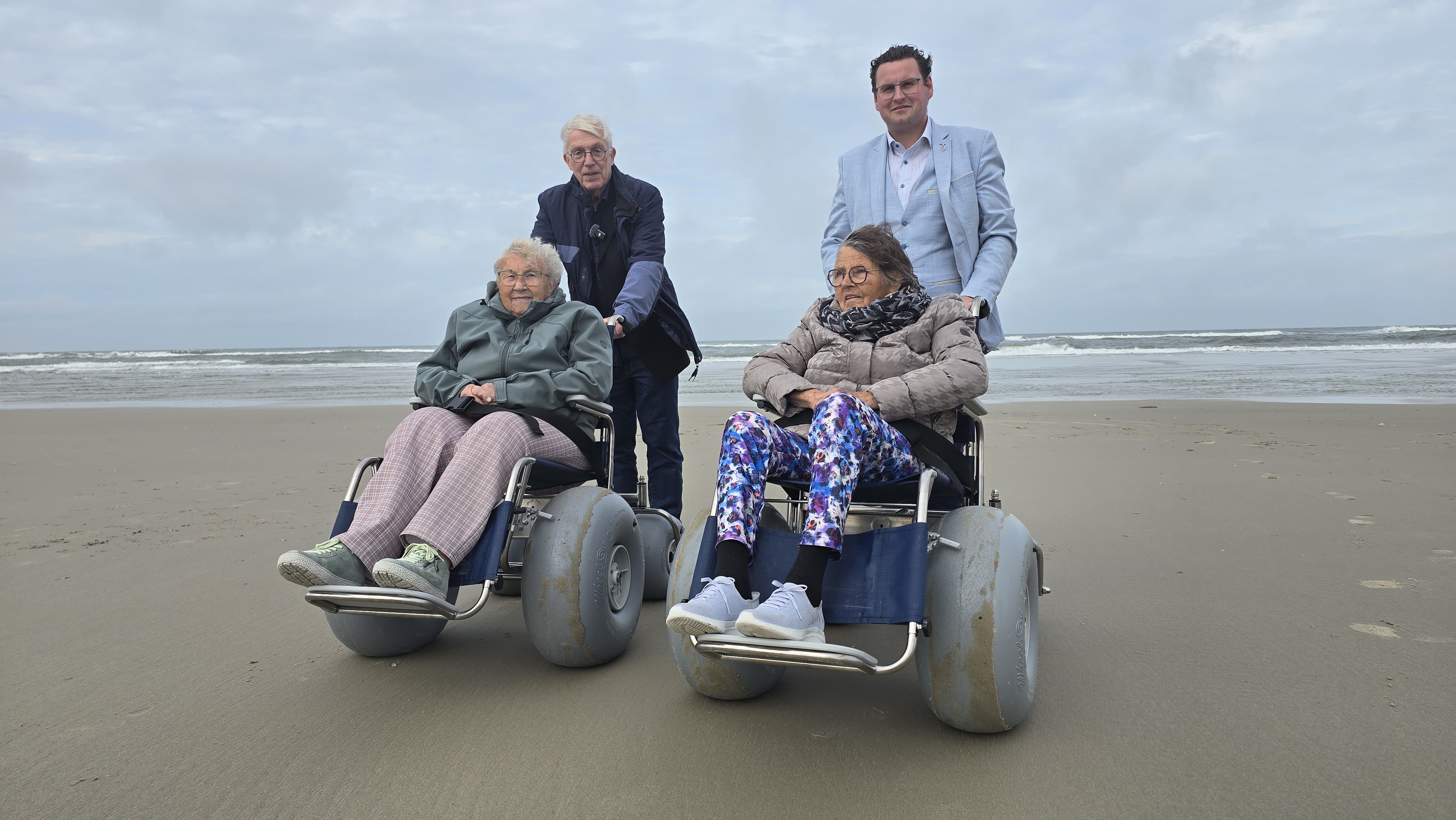 Twee vrouwen zitten in een strandrolstoel en worden geduwd door twee mannen.