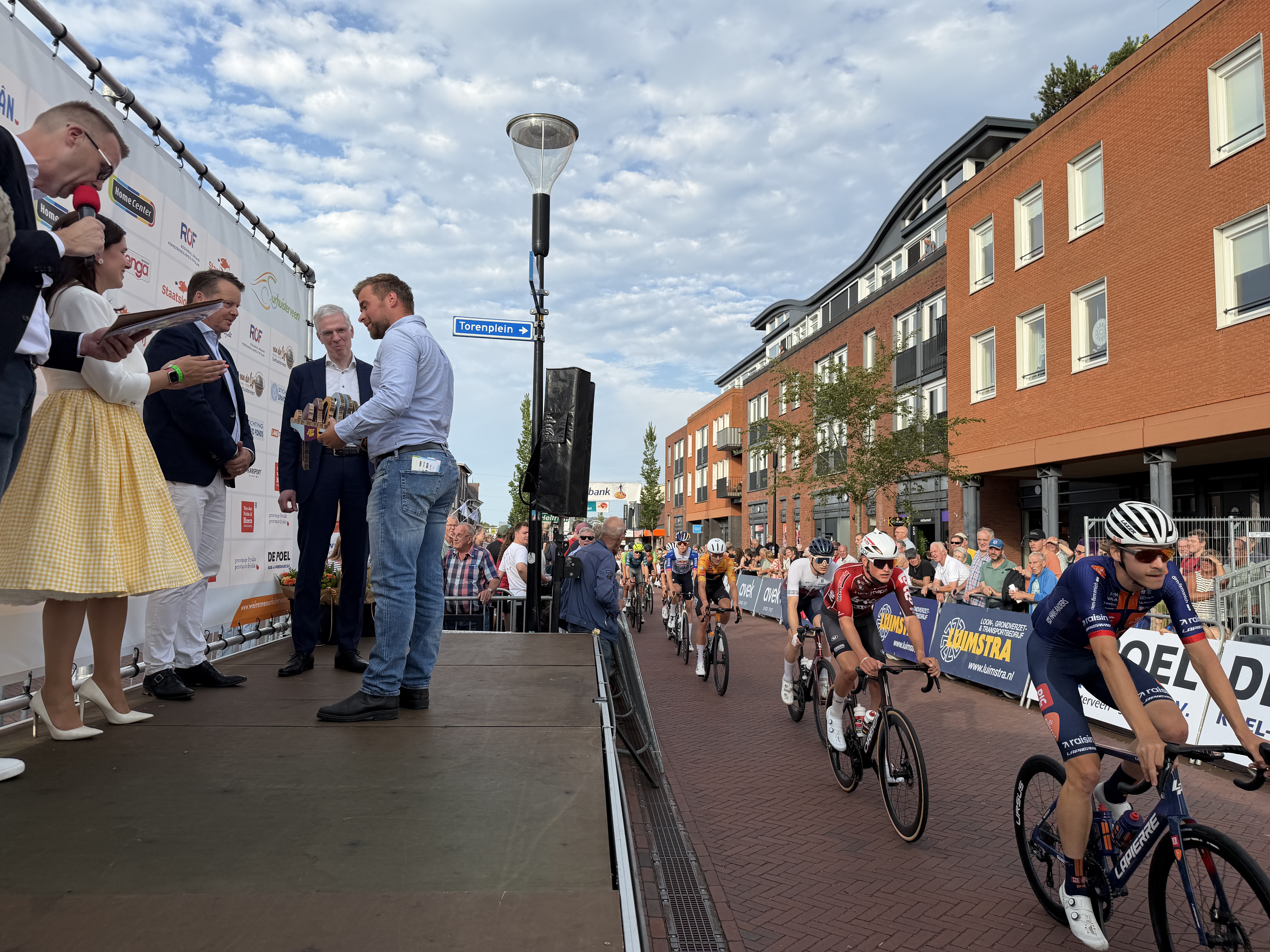 Aan de linkerkant wordt de helm als symbool voor het Doortrapseizoen overgedragen. Aan de rechterkant fietsen wielrenners over de weg.