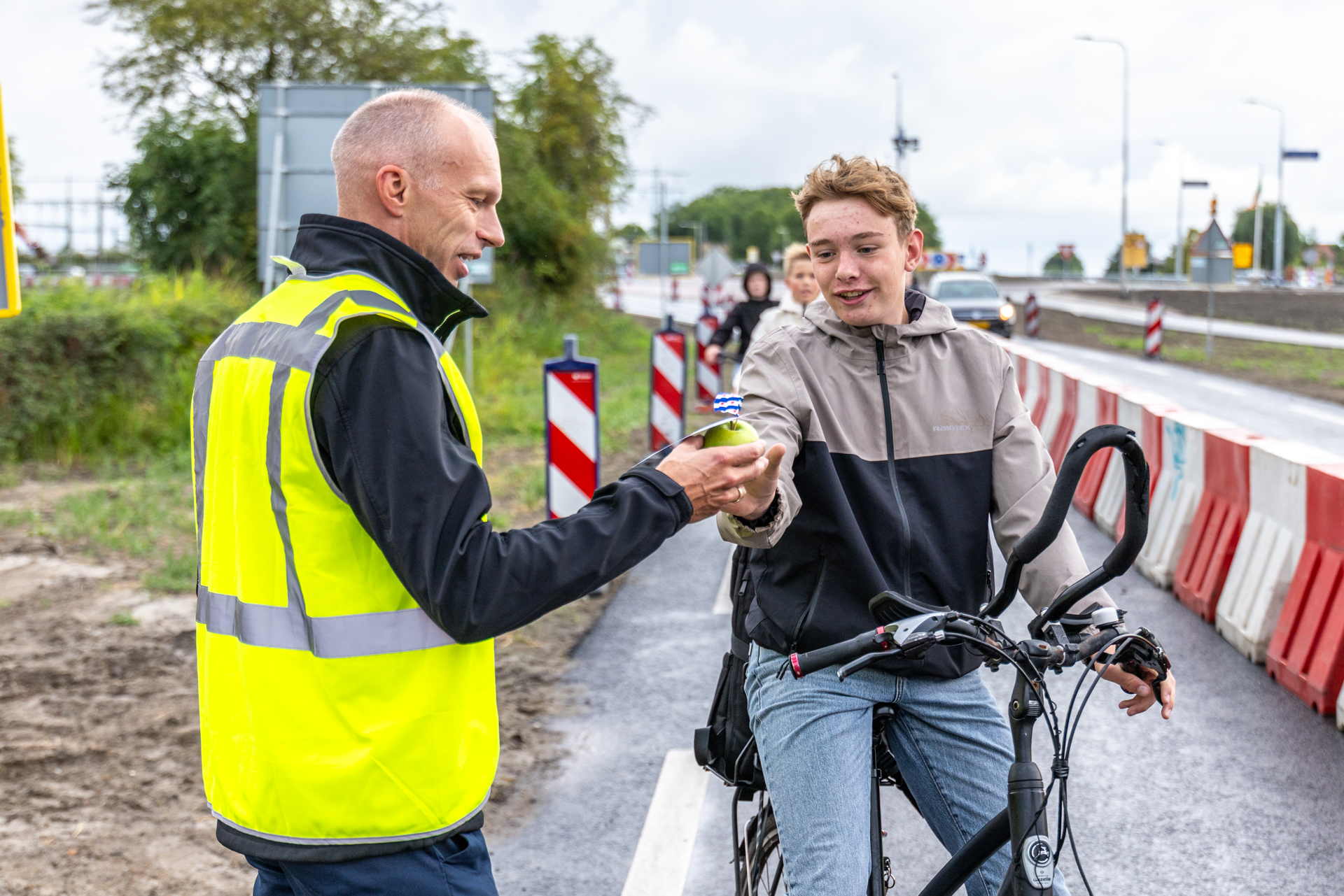 Een man in een geel hesje deelt een appel uit aan een jongen op de fiets.