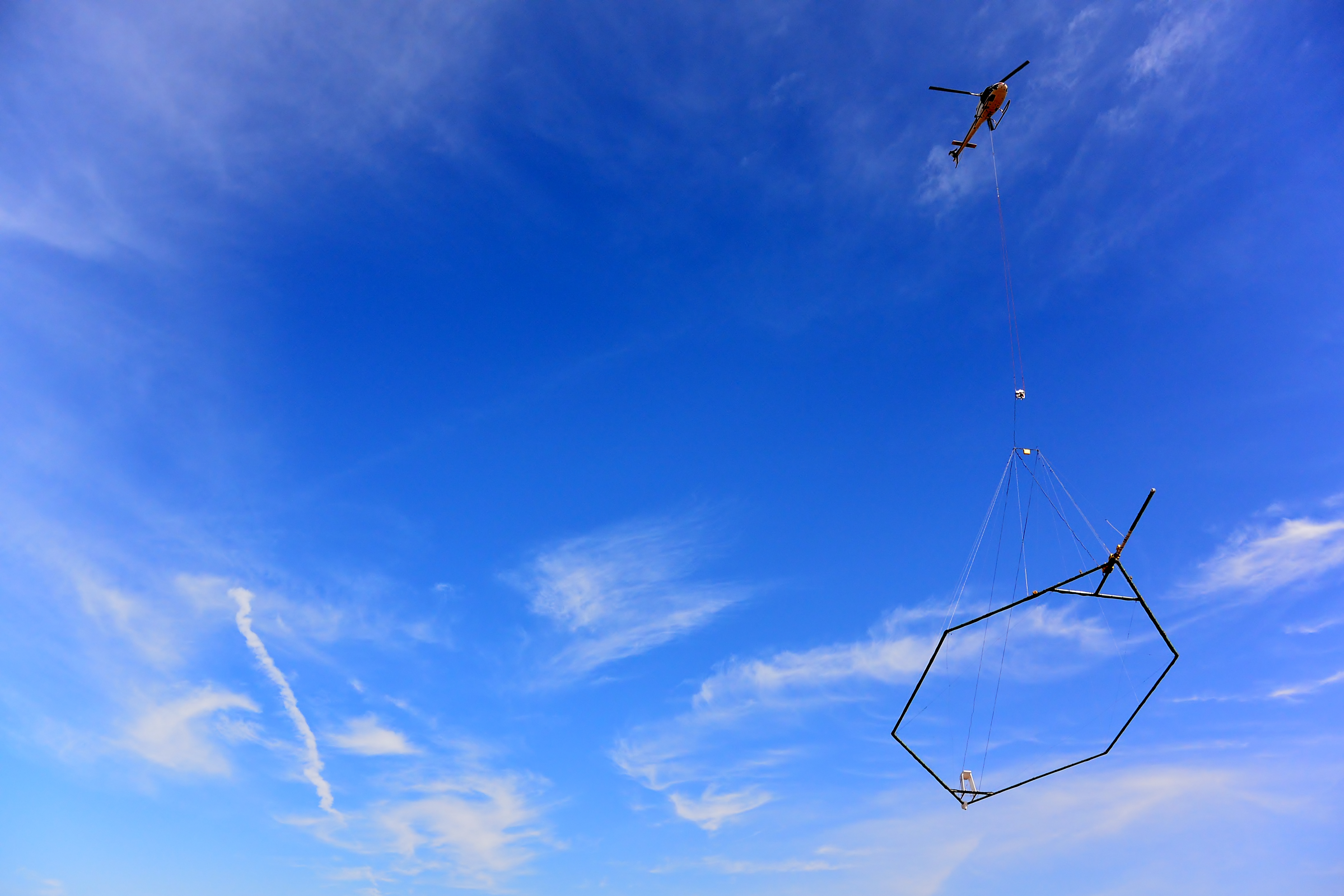 Een bijna strakblauwe lucht waar een helikopter in vliegt waar een groot scanapparaat onder hangt.
