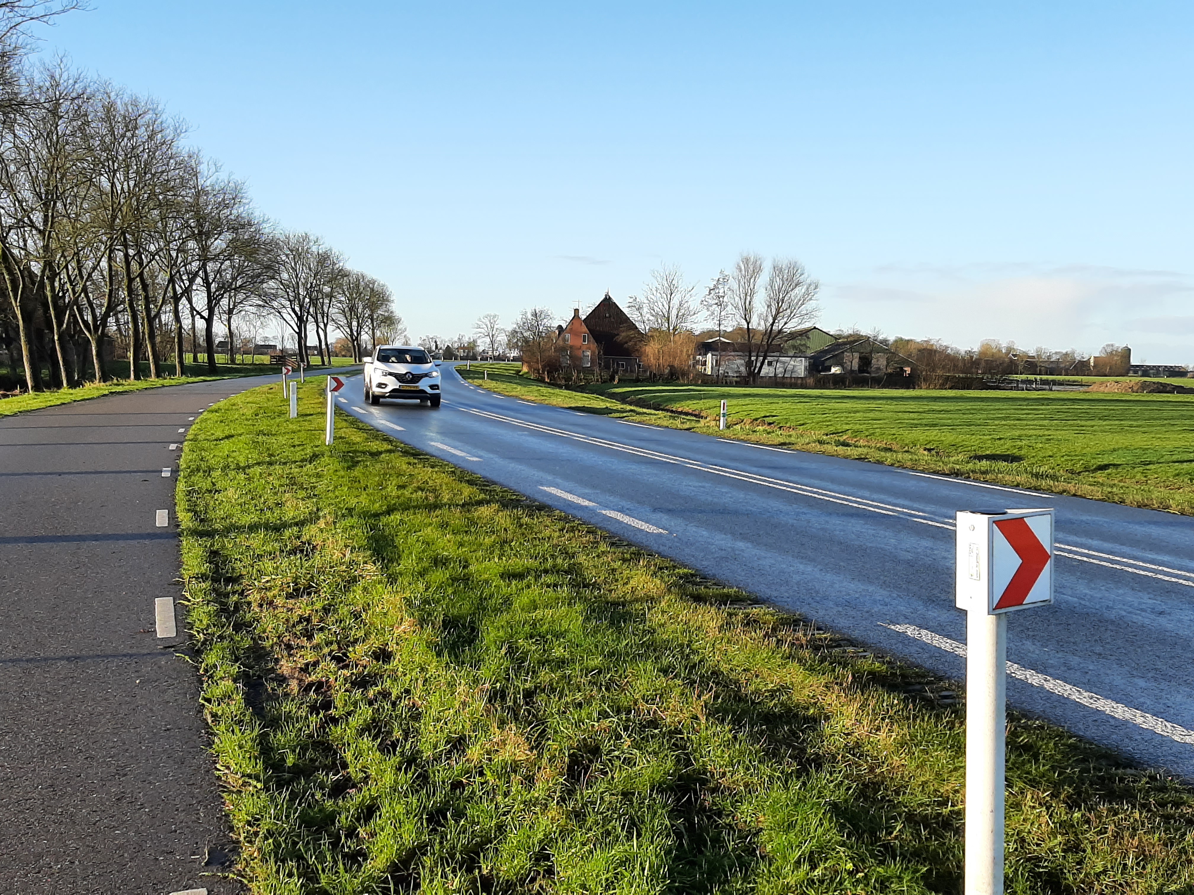 Een weg waar een auto op rijdt met berm en een boerderij op de achtergrond
