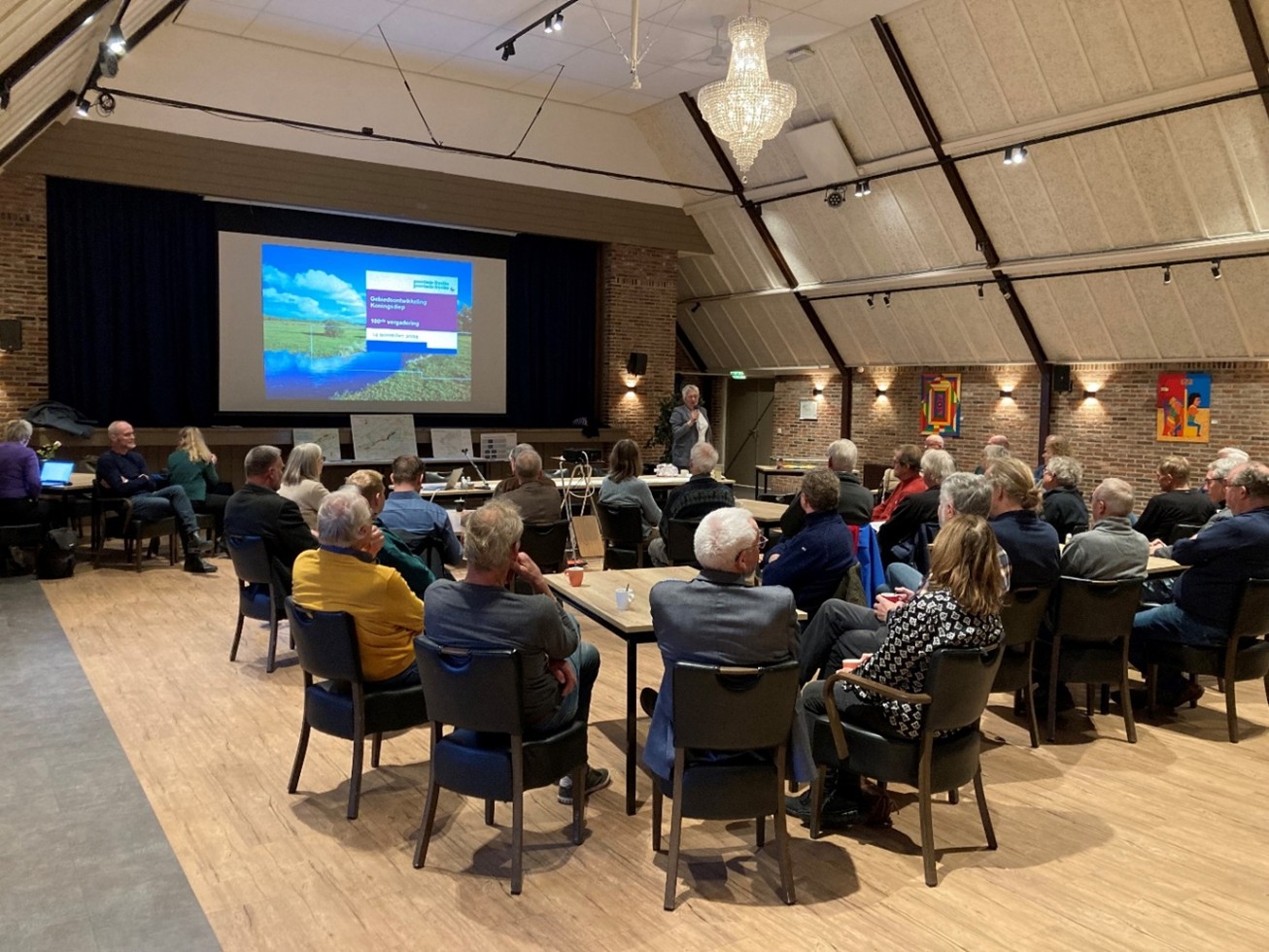 Een groep mensen zit aan tafeltjes te luisteren naar een spreker die bij een groot scherm staat.