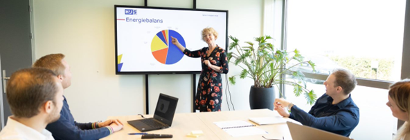 Een vrouw staat voor een presentatiescherm en er zitten 4 personen aan tafel te luisteren.