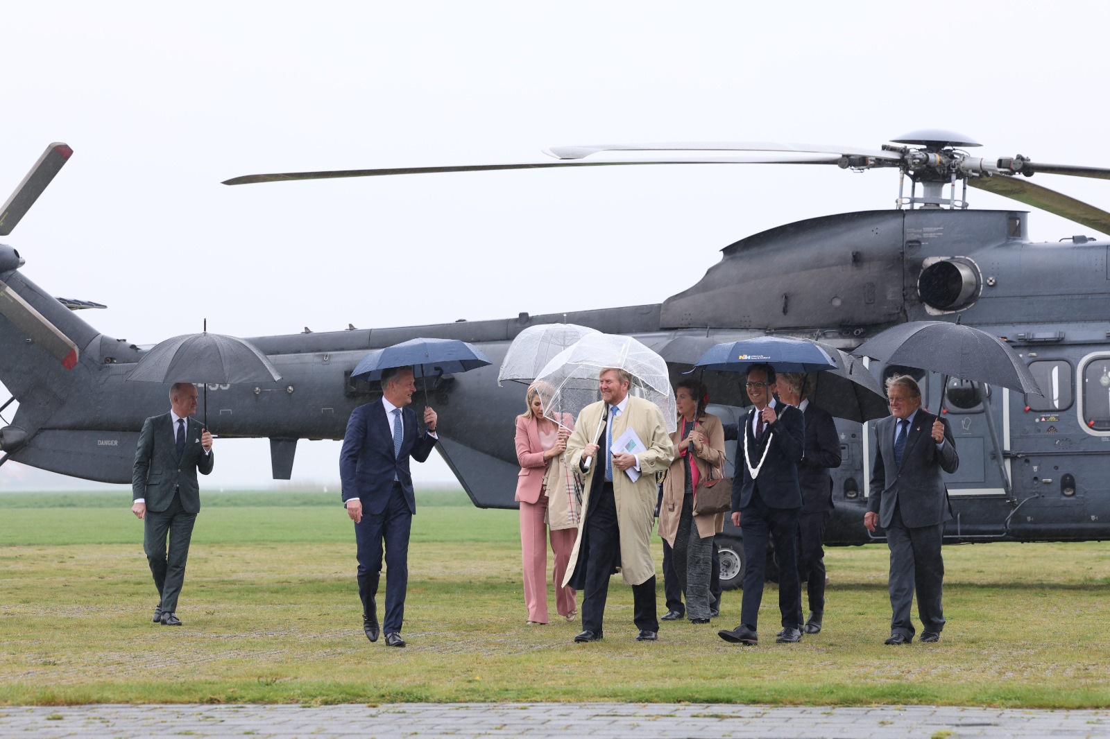 Het Koningspaar stapt uit de helicopter met paraplu's en worden ontvangen door burgemeesters en commissarissen van de Koning
