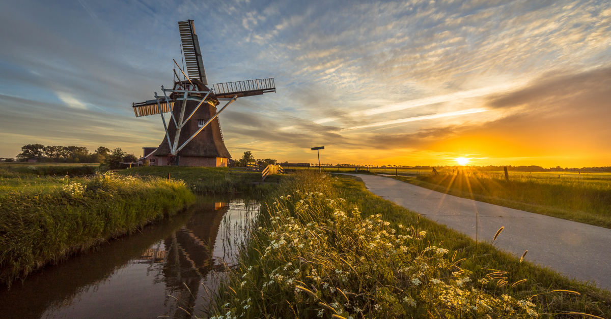 Een molen staat in een weiland waar een sloot en een pad te zien is. De zon gaat onder.