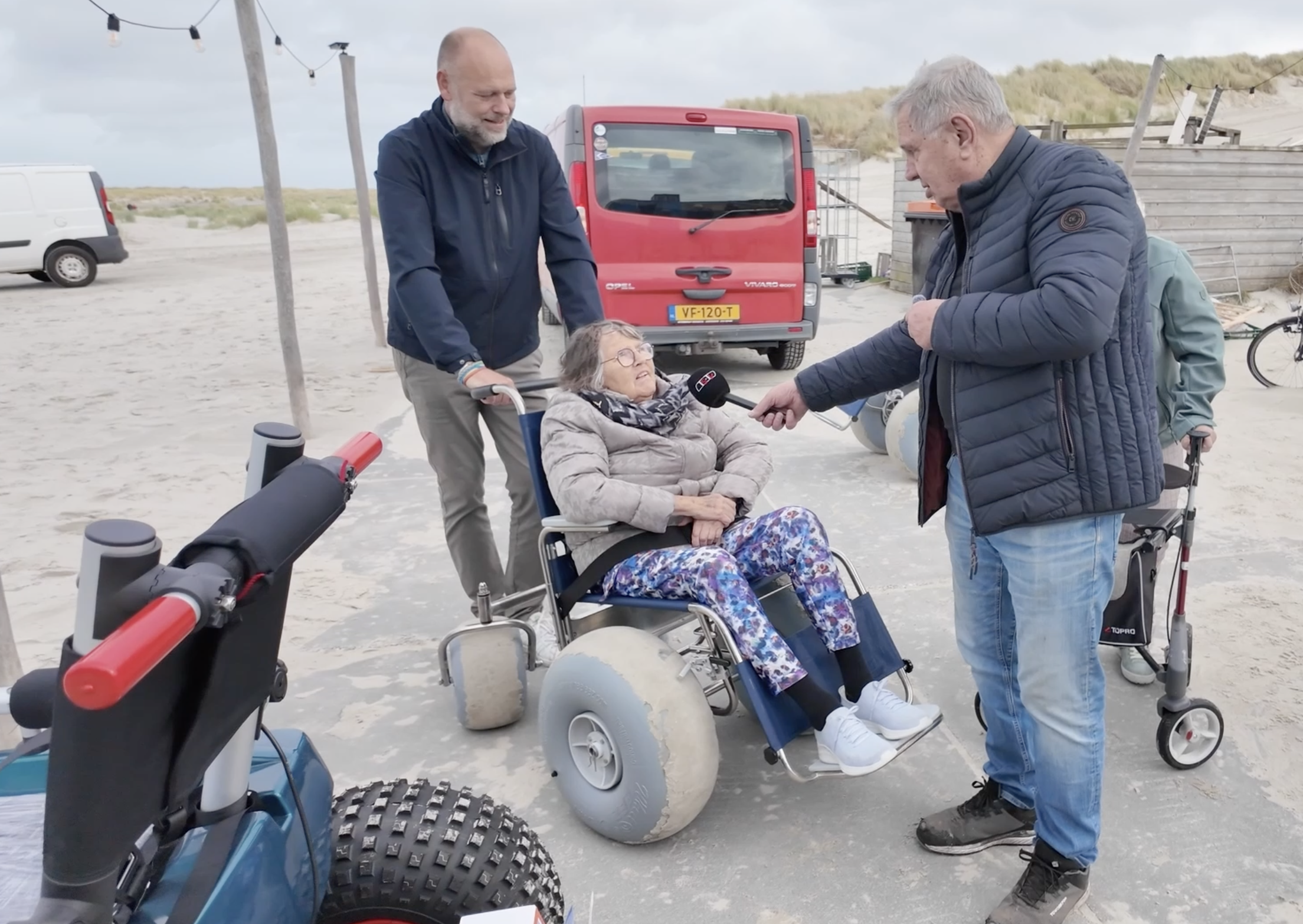 Een oudere vrouw in een nieuwe strandrolstoel wordt geïnterviewd door een reporter van Terschelling TV.