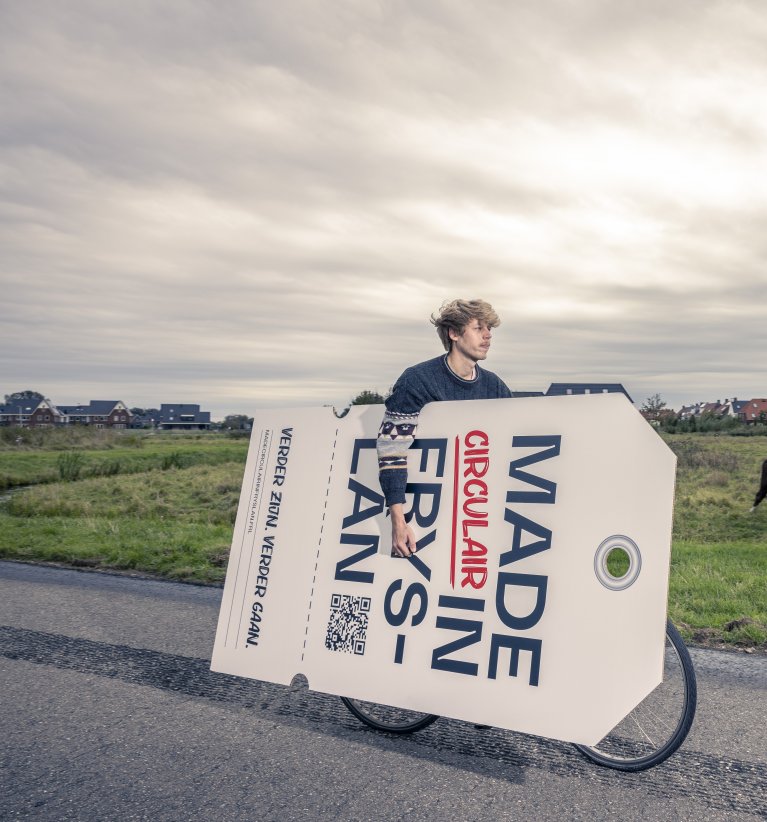 Jongeman op een fiets met een enorm groot label onder zijn arm waar op staat: Made in circulair Fryslân.
