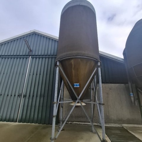 Een silo staat voort een stal op een betonnen vloer met een rooster in de grond.