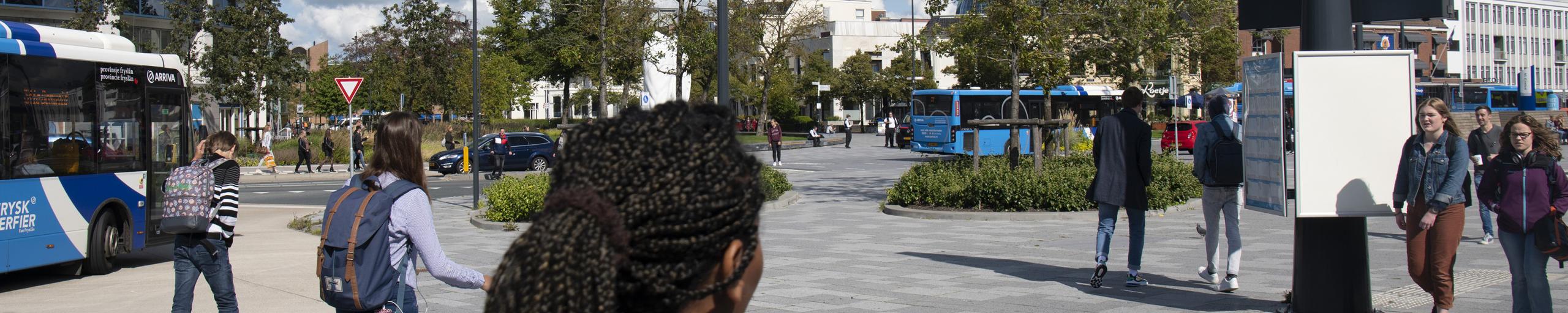Een vrouw op de voorgrond met op links twee bussen die het busstation afrijden
