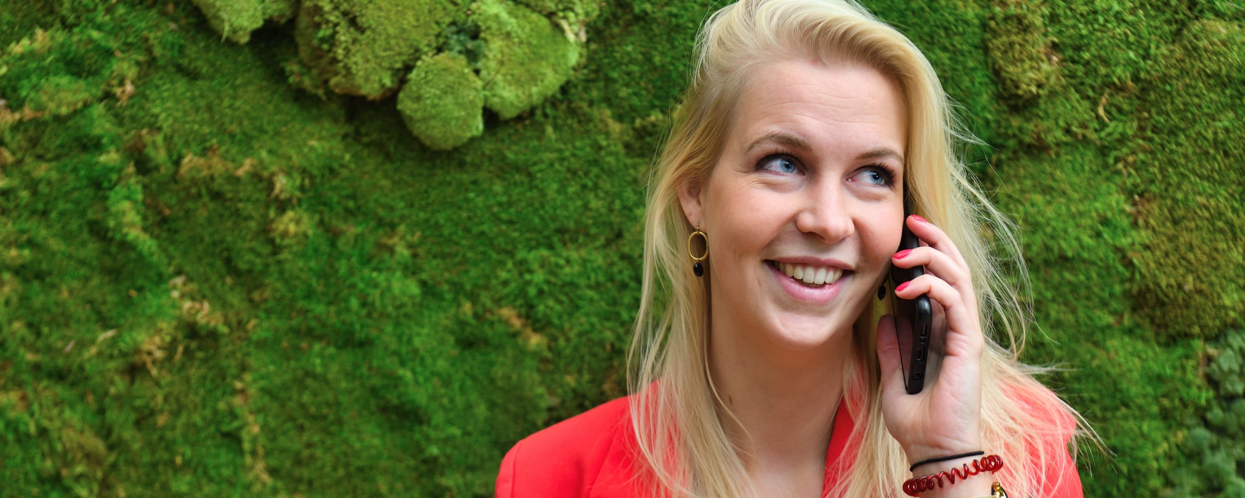 Een blonde vrouw met een telefoon aan haar oor voor een bemoste achtergrond.