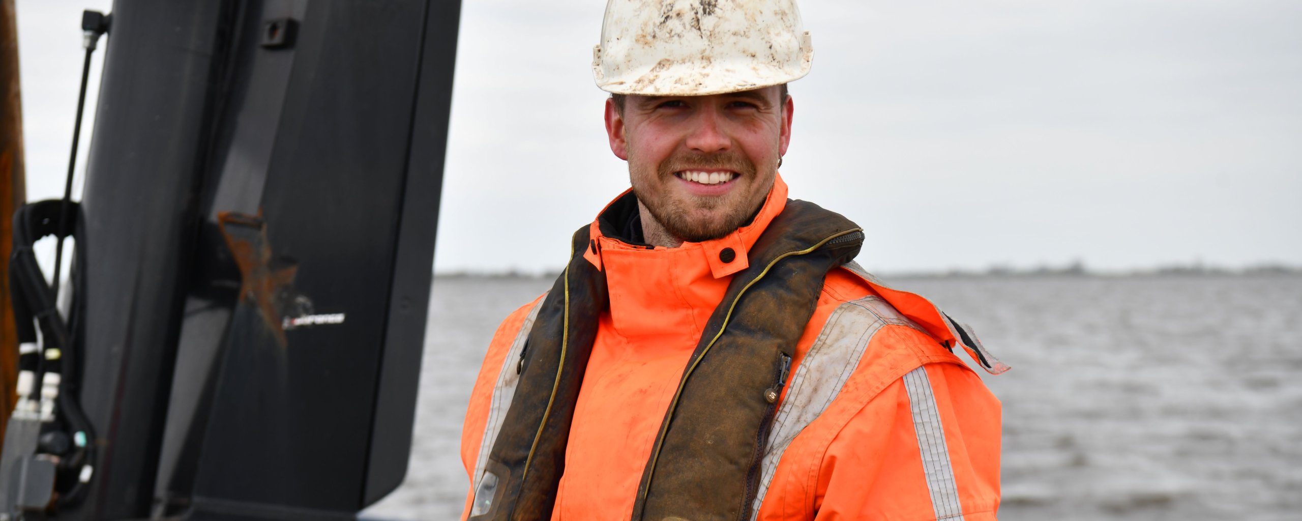 Een man in een oranje verkeersjas en een helm op zit in een schip met water op de achtergrond.