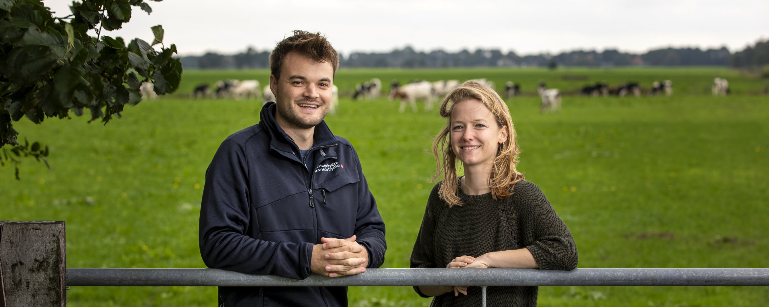 Een man en een vrouw staan tegen een hek aangeleund met op de achtergrond koeien in de wei.