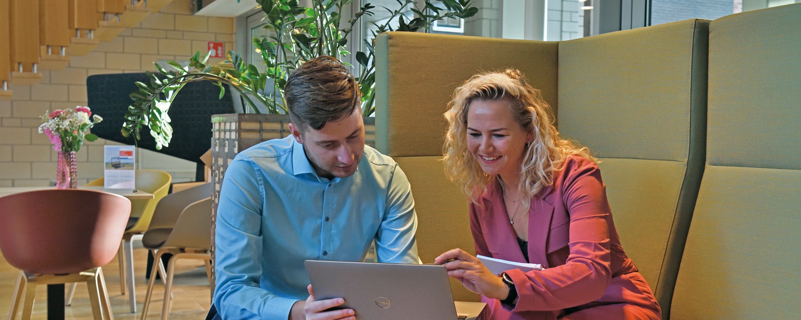 Twee personen zitten op een bank in de publiekshal van het provinciehuis naar een laptopscherm te kijken